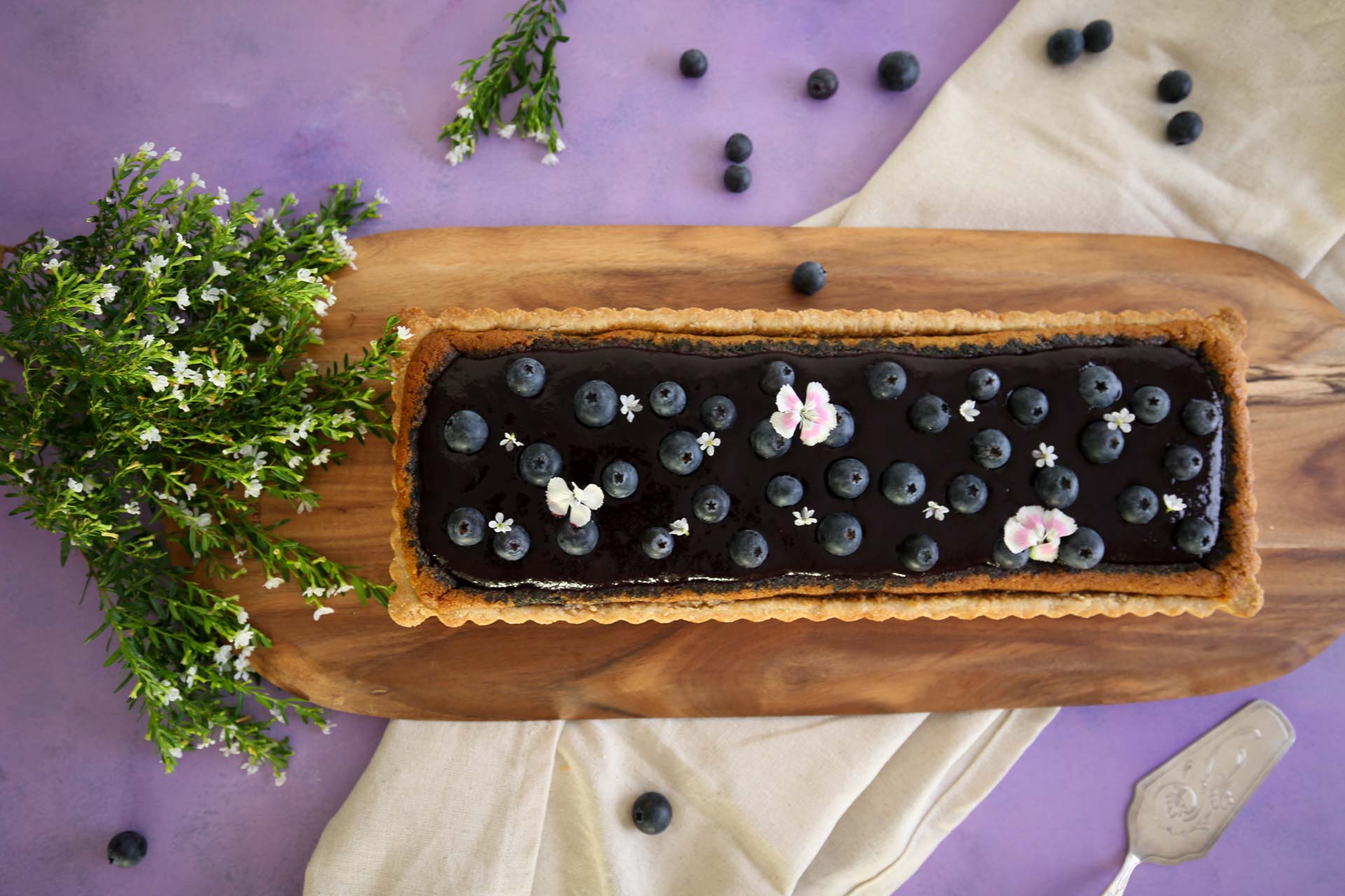 Top view of a blueberry tart on a wooden board decorated with extra blueberries and flowers